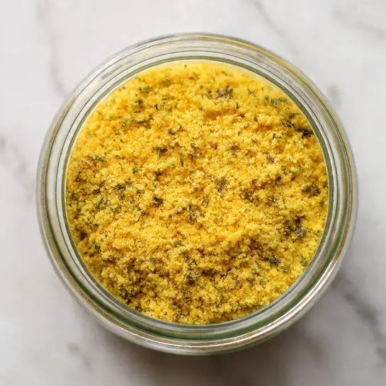 A close-up image showing a clear glass jar filled with finely ground yellow seasoning. The seasoning has a slightly grainy texture with small bits of herbs and spices visible throughout. The jar is viewed from above, centered in the frame, placed on a white marbled surface. The yellow color of the seasoning is bright and uniform, covering the entire visible surface inside the jar. Photo taken with an iphone --ar 4:5 --v 7