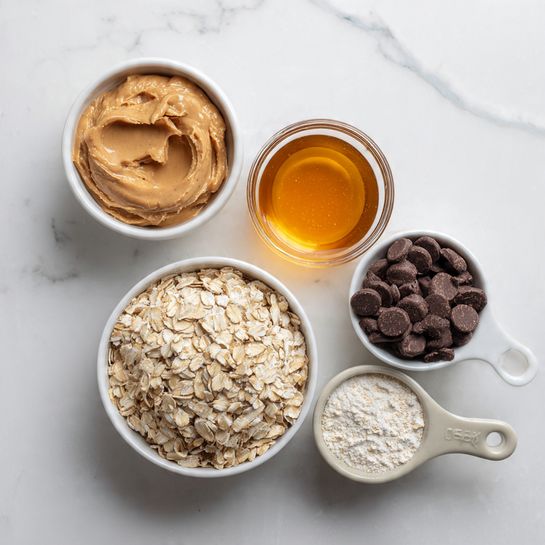 The image shows five small containers neatly arranged on a white marbled surface. On the top left, there is a white bowl filled with smooth, light brown peanut butter. To its right, a clear glass bowl holds a shiny, golden amber liquid, likely honey. Below the peanut butter bowl is another white bowl filled with pale beige rolled oats that have a rough texture. Positioned to the right of the oats are two measuring cups with light gray handles; the larger one contains small, round dark brown chocolate pieces, while the smaller one holds off-white powdered flour. The containers are closely placed, showcasing contrasting textures and natural colors. photo taken with an iphone --ar 4:5 --v 7