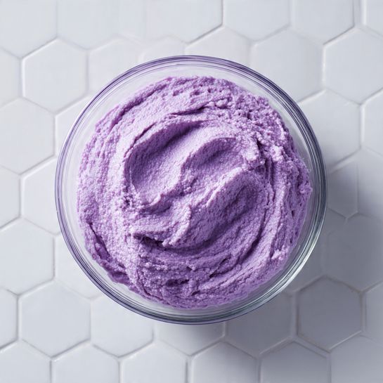 A close-up top view of a clear glass bowl filled with a thick, creamy mixture that is a purple color with a slightly grainy texture. The mixture looks smooth but dense, with some peaks and swirls created by stirring. The bowl is placed on a white marbled surface with a subtle hexagonal tile pattern in the background. The photo taken with an iphone --ar 4:5 --v 7