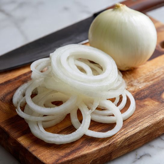 A close-up of thin, white onion rings neatly stacked in a small pile on a wooden cutting board with a brown grain pattern. Behind the rings sits a peeled whole onion with a smooth, creamy white surface and a rounded shape. Part of a chef’s knife blade is visible in the background, lying flat on the same wooden board. The photo is taken on a white marbled surface. Photo taken with an iphone --ar 4:5 --v 7