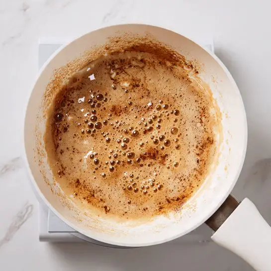 The image shows a close-up of a white pot with a brown bubbling liquid inside. The liquid has a foamy, frothy texture with many bubbles rising to the surface, indicating it is boiling. The inside walls of the pot have splashes of the same brown liquid. The pot is placed on a stovetop burner. The overall scene has a white marbled surface background. photo taken with an iphone --ar 4:5 --v 7