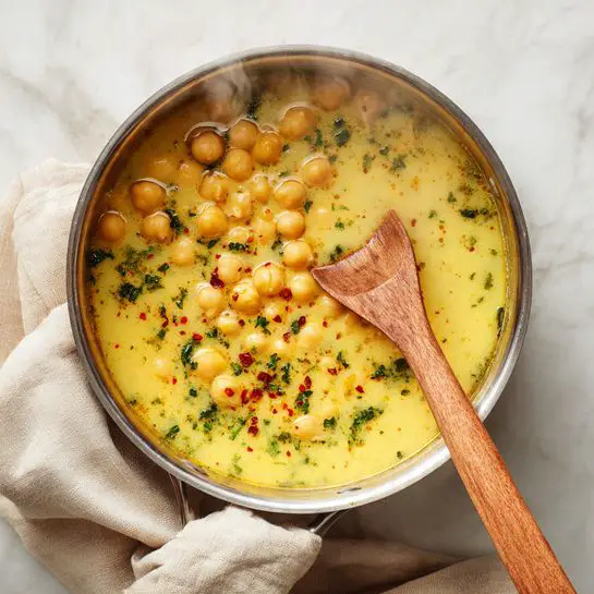 A close-up view of a metal pot filled with creamy chickpea soup, with a light yellow, smooth broth holding round chickpeas floating on top. The soup has a sprinkle of green herbs and small bits of red spice, adding color contrast on the surface. A wooden spoon is dipped into the soup on the right side of the pot, with steam rising gently from the hot liquid. The pot rests on a white marbled surface, and a soft beige cloth is draped over the edge of the pot. Photo taken with an iphone --ar 4:5 --v 7