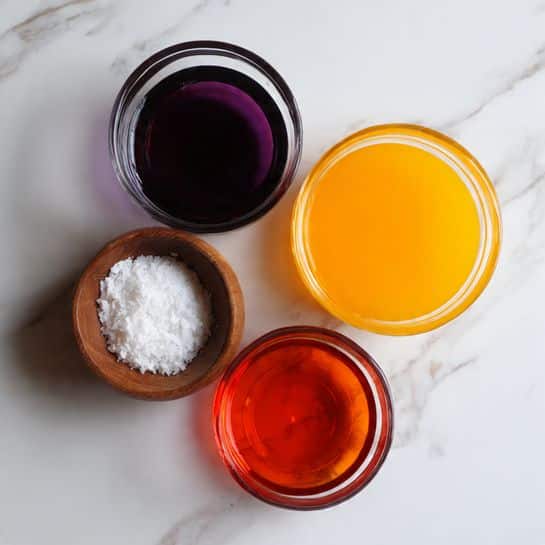 The image shows a top view of three clear glass bowls filled with different colored liquids arranged on a white marbled surface. The bowl at the top left contains a dark purple liquid, the bowl at the top right holds a bright orange liquid, and the bowl at the bottom left has a vibrant red liquid. Next to the vibrant red bowl is a small wooden bowl filled with a white granular substance. The arrangement is simple with a clean and bright look. photo taken with an iphone --ar 4:5 --v 7