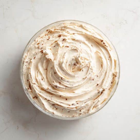 A close-up view of a round clear bowl filled with a single thick layer of creamy, light beige spread with small darker bits mixed in. The surface has visible swirls and soft peaks, creating a textured, smooth appearance. The bowl sits on a white marbled surface. photo taken with an iphone --ar 4:5 --v 7