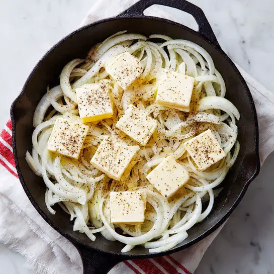 A black cast iron pan filled with many thin white onion slices spread out evenly across the pan. On top of the onions, there are six small square pats of pale yellow butter, each sprinkled lightly with black pepper, resting on different spots over the onions. The pan sits on a white marbled surface with a white cloth with red stripes partially visible beneath it. Photo taken with an iphone --ar 4:5 --v 7