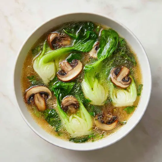 This is a white bowl filled with a clear golden broth, showing some light bubbles on the surface. Inside the broth, there are several layers of fresh green bok choy with pale white stems, arranged evenly in the bowl. Scattered among the bok choy are thin slices of brown shiitake mushrooms with a slightly curved shape, floating gently in the broth. The bowl is placed on a white marbled surface. Photo taken with an iphone --ar 4:5 --v 7