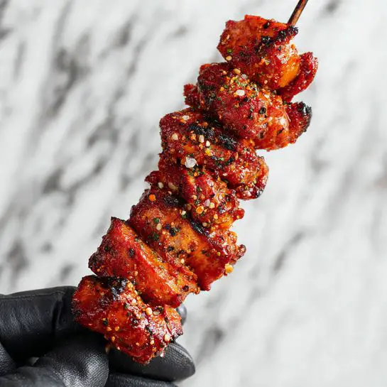 The image shows a skewer with six pieces of grilled meat stacked closely together. Each piece is coated with a reddish-orange spice mix that has visible black pepper flakes. The meat looks tender and slightly shiny with a lightly charred texture on the edges. The skewer is held by a gloved woman's hand with a black glove, and the background is a white marbled surface. photo taken with an iphone --ar 4:5 --v 7