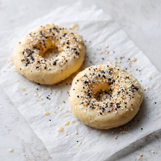 The image shows two raw dough bagels on a piece of parchment paper placed on a white marbled surface. Each bagel is round with a hole in the middle, and they are topped with white sesame seeds and black poppy seeds scattered evenly across their slightly wet, pale dough surface. The texture of the dough looks soft and moist, ready to be baked. The parchment paper has small droplets and seeds around the bagels, adding a sense of freshness and preparation. photo taken with an iphone --ar 4:5 --v 7