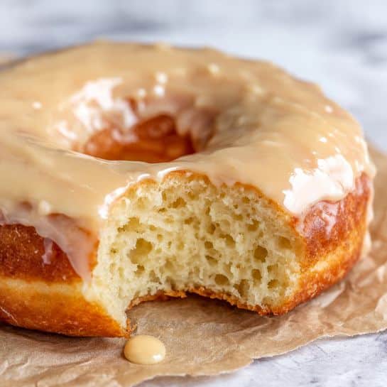 A close-up view of a donut with one big bite showing the fluffy, soft inside texture with small holes. The donut is thick and golden brown on the outside. On top, there is one thick layer of shiny, smooth beige icing that covers the whole top surface. The donut is placed on crinkled light brown parchment paper with a small drop of icing visible near it, against a white marbled background. Photo taken with an iphone --ar 4:5 --v 7
