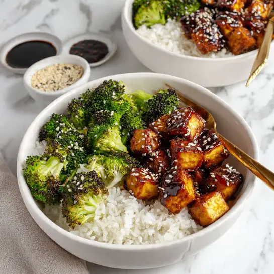 A white bowl on a white marbled surface holds a colorful meal with three layers: at the bottom is a bed of plain white rice, on the left side bright green broccoli florets with some parts slightly charred, and on the right side dark brown glazed tofu cubes sprinkled with white sesame seeds creating a glossy texture. A gold fork rests inside the bowl on the right side, partially under the tofu. Behind this bowl, a second white bowl with a similar meal is partially visible. Near the bowls, two small white dishes contain black sauce and sesame seeds mix. Photo taken with an iphone --ar 4:5 --v 7