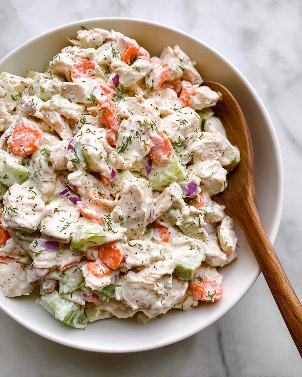 The image shows a white bowl filled with a creamy chicken salad, mixed with pieces of cooked white chicken, pale green celery chunks, and slices of orange carrot. The salad is coated in a white creamy dressing with visible specks of black pepper and green herbs scattered throughout. Small bits of purple onion are also mixed in, adding more color contrast. A wooden spoon is resting in the bowl on the right side, slightly covering some of the salad. The bowl is placed on a white marbled surface photo taken with an iphone --ar 4:5 --v 7