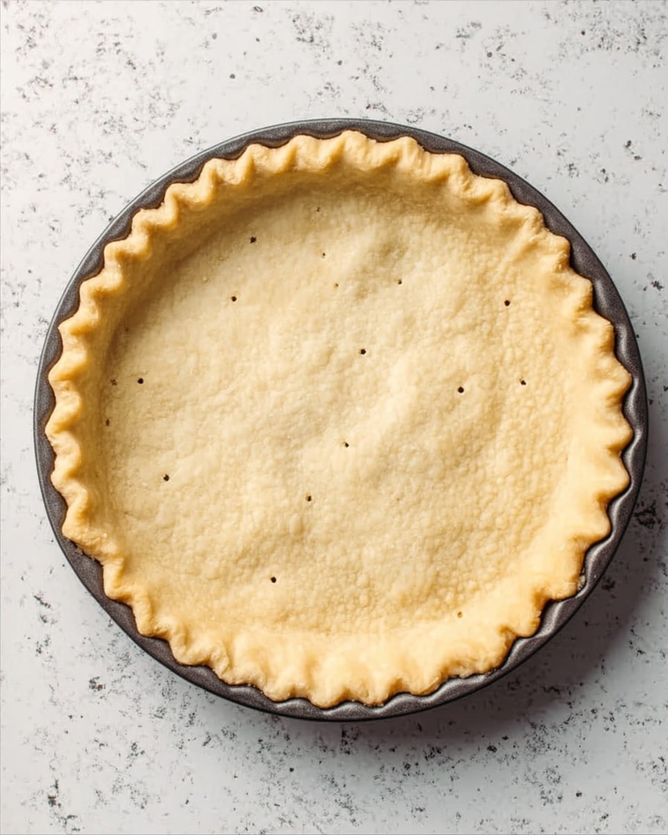 A single-layer light golden pie crust with a slightly crimped edge sits in a dark metal pie pan. The crust has small evenly spaced fork holes in the bottom center. The pie pan rests on a white marbled surface. photo taken with an iphone --ar 4:5 --v 7