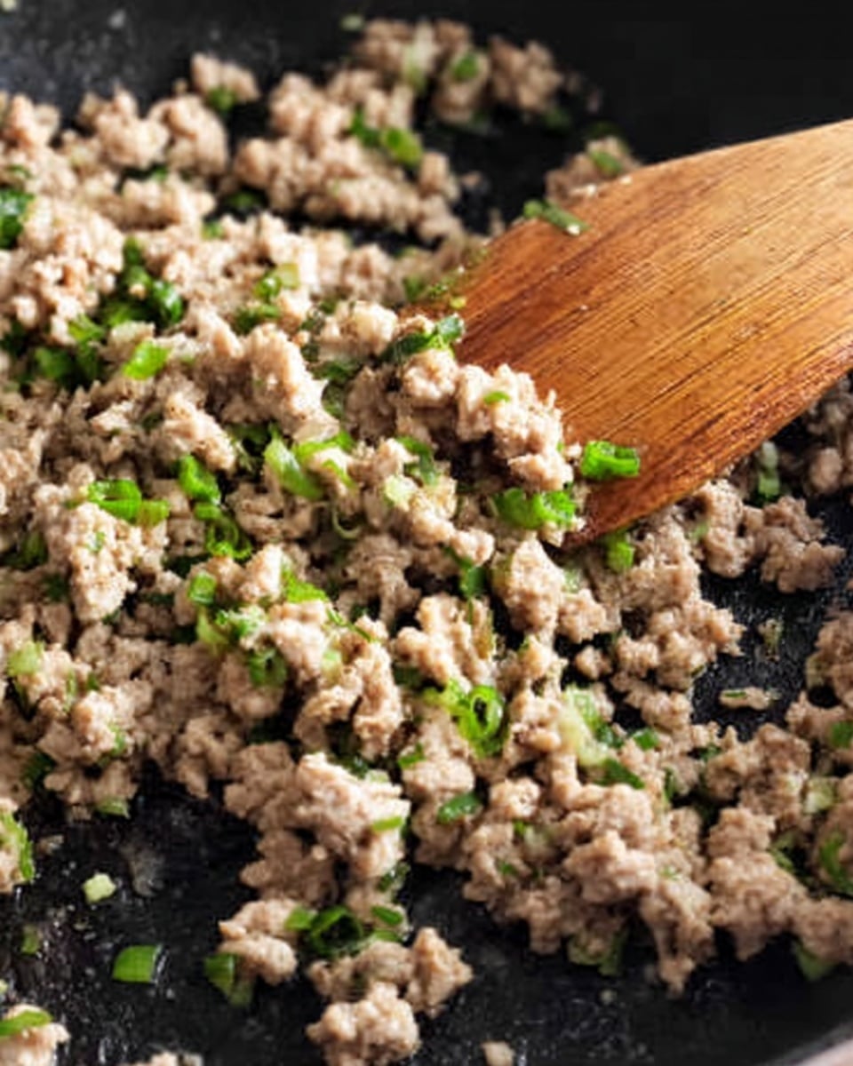 The image shows a close-up of cooked ground meat mixed with small pieces of green onions, all in a black pan. A wooden spatula with a smooth texture lies on top of the meat, slightly lifting some of it. The meat is light brown and crumbly in texture, with bits of green onion scattered throughout, adding small bright green spots to the dish. The cooking pan's surface is dark with some slight shine from oil or moisture. photo taken with an iphone --ar 4:5 --v 7