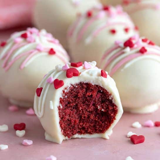 The image shows round cake balls with two layers: a deep red, moist cake inside and a smooth white coating outside. The white layer is decorated with thin swirls and small red, pink, and white heart-shaped sprinkles on top and around the cake balls. One cake ball in the front is bitten into, revealing the rich red inside. The treats are placed on a pink surface with a soft blurred background showing more similar cake balls. photo taken with an iphone --ar 4:5 --v 7