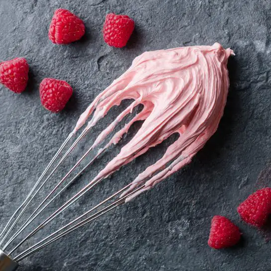 The image shows a white mixing beater with thick, smooth pink frosting swirled around its metal blades, resting on a dark textured surface. Nearby, there are several fresh raspberries scattered loosely around the beater, their red color bright and contrasting with the pale pink frosting. The scene is simple, focusing on the textures of the creamy frosting and the bumpy surfaces of the raspberries. photo taken with an iphone --ar 4:5 --v 7