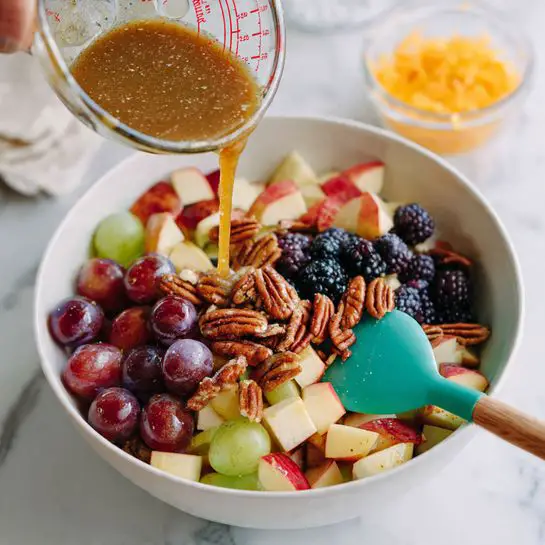 The image shows a white bowl filled with a colorful mix of chopped apples, red grapes, blackberries, and chopped pecans, all coated in a light brown dressing. A turquoise spatula rests inside the bowl on the right side. The fruit and nuts create layers of deep purple, red, light beige, and golden brown colors. In the background, a hand is grating orange zest from an orange over a white marbled surface. Another part shows a clear glass measuring cup with a dark brown dressing being stirred, and the dressing is poured over the fruit mix in the white bowl. The overall scene is brightly lit on a white marbled surface with a fresh and natural look photo taken with an iphone --ar 4:5 --v 7