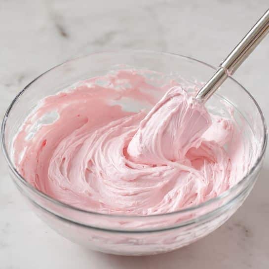 A clear glass bowl with a spout filled with smooth, thick pink frosting that has been stirred, showing swirled textures and soft peaks. A metal spatula is dipped into the frosting, lifting a swirl of the creamy pink mixture above the bowl. The surface beneath the bowl is a white marbled texture. photo taken with an iphone --ar 4:5 --v 7