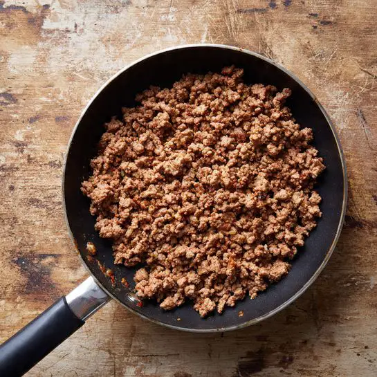 A black frying pan with a long black and silver handle is filled with cooked ground meat that is brown and crumbly. The meat is spread evenly inside the pan, showing a textured surface with small browned bits throughout. The pan sits on a wooden surface with worn patches and a rustic look, but the background should be a white marbled texture. photo taken with an iphone --ar 4:5 --v 7