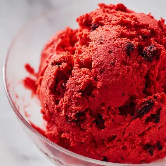 The image shows a close-up of a single large clump of red dough with a rough, uneven texture inside a clear glass mixing bowl. The dough is bright red with some darker red and slightly grayish spots. The bowl is placed on a white marbled surface. Part of a white kitchen appliance with a bit of black is visible on the right side in the background. The photo taken with an iphone --ar 4:5 --v 7