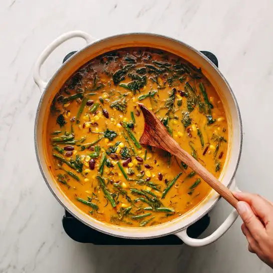 A large white pot filled with a creamy, warm orange soup with visible pieces of green vegetables, yellow corn, and some dark beans or seeds mixed evenly throughout. A woman's hand holding a wooden spoon is stirring the soup, creating a slight ripple effect on the surface. The pot sits on a black hot plate, and the background is a white marbled texture. photo taken with an iphone --ar 4:5 --v 7