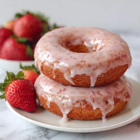 Two pink donuts stacked on top of each other sit on a white plate, each donut covered with a shiny, light pink glaze that drips slightly over the sides. The donuts have a soft, slightly rough texture with bits of darker pink visible inside. In the background, fresh whole strawberries with green leaves add a bright red contrast, all set on a white marbled surface. Photo taken with an iphone --ar 4:5 --v 7