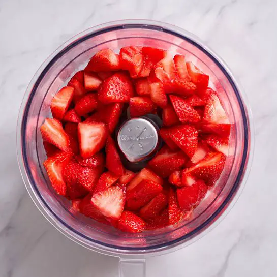 A clear blender container filled with small, bright red chopped pieces of strawberries. The sharp silver blades are centered inside the container, surrounded by the strawberry pieces which cover the bottom layer. The background is a white marbled surface reflected faintly on the clear sides of the blender container. Photo taken with an iphone --ar 4:5 --v 7