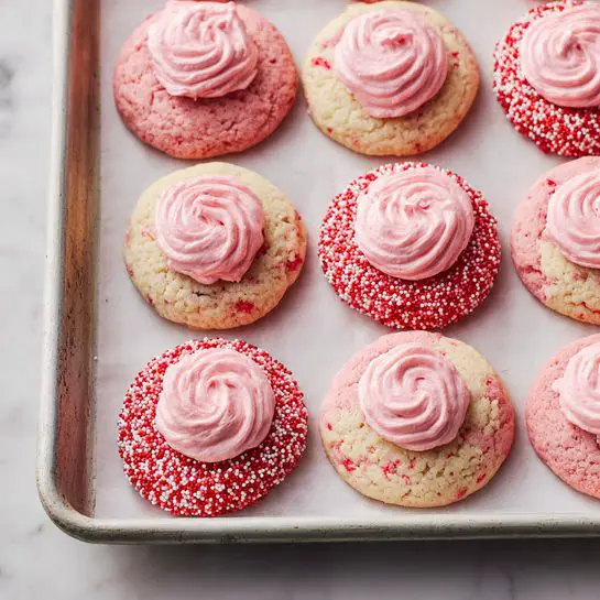 The image shows a baking tray with 12 round cookies arranged in neat rows on white parchment paper. Each cookie has two layers: the bottom layer is a soft, light pink round base with a slightly cracked texture and small colorful specks inside, and the top layer is a dollop of smooth pink frosting placed in the center. Some cookies have their edges covered in small round sprinkles in shades of red, white, and pink, giving them a bumpy texture along the rim, while others have plain edges. The baking tray sits on a white marbled surface. Photo taken with an iphone --ar 4:5 --v 7