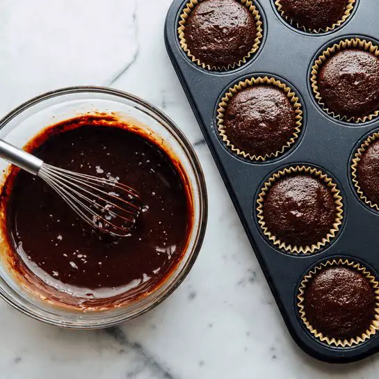 The image shows a white plate with eight dark chocolate cupcakes arranged close together. Each cupcake has a shiny, thick layer of smooth dark chocolate frosting on top, with a small strawberry heart slice placed in the center of the frosting. One cupcake has a bite taken out, revealing a soft, light pink strawberry cream filling inside the dark chocolate cake. Three large chocolate-covered strawberries with green tops sit on the white marbled surface near the plate. The lighting highlights the glossy frosting and fresh strawberries. photo taken with an iphone --ar 4:5 --v 7