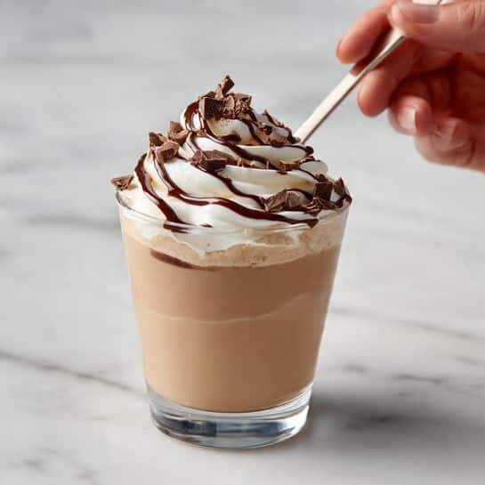 A clear glass cup filled with a light brown, creamy drink topped with a generous swirl of white whipped cream. On top of the whipped cream, there are drizzles of dark chocolate syrup creating thin lines that flow down slightly. Small chocolate chunks or chips are scattered on the whipped cream as well. The glass is set on a white marbled surface with a woman's hand holding a light-colored spoon inside the drink. Photo taken with an iphone --ar 4:5 --v 7