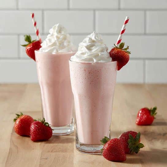 Two tall clear glasses hold strawberry milkshakes with a light pink color and slightly foamy texture at the bottom. Each milkshake is topped with a large swirl of white whipped cream that looks soft and fluffy. A red and white striped straw is placed on the right side of each glass, leaning slightly outwards. Near the top edge of the front glass, there is a fresh whole red strawberry with its green leaves still attached. Around the glasses on the light wooden surface are several more whole strawberries, with one visible in the front right corner. The background shows a white marbled surface and white subway tiles, giving a clean and bright look. photo taken with an iphone --ar 4:5 --v 7