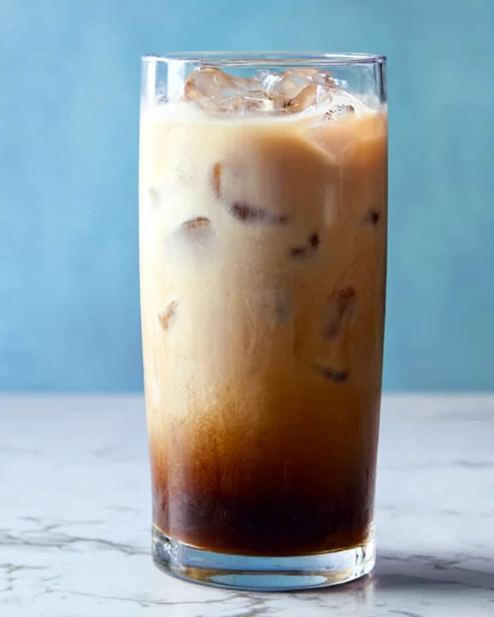 The image shows a tall clear glass filled with an iced drink that has three main layers. The bottom layer is dark brown, appearing dense and rich. The middle layer is lighter brown, blending softly into the bottom. The top layer is creamy white with ice cubes floating and slightly melting, creating a smooth texture. The background is a soft blue color, and the whole scene rests on a white marbled surface. photo taken with an iphone --ar 4:5 --v 7