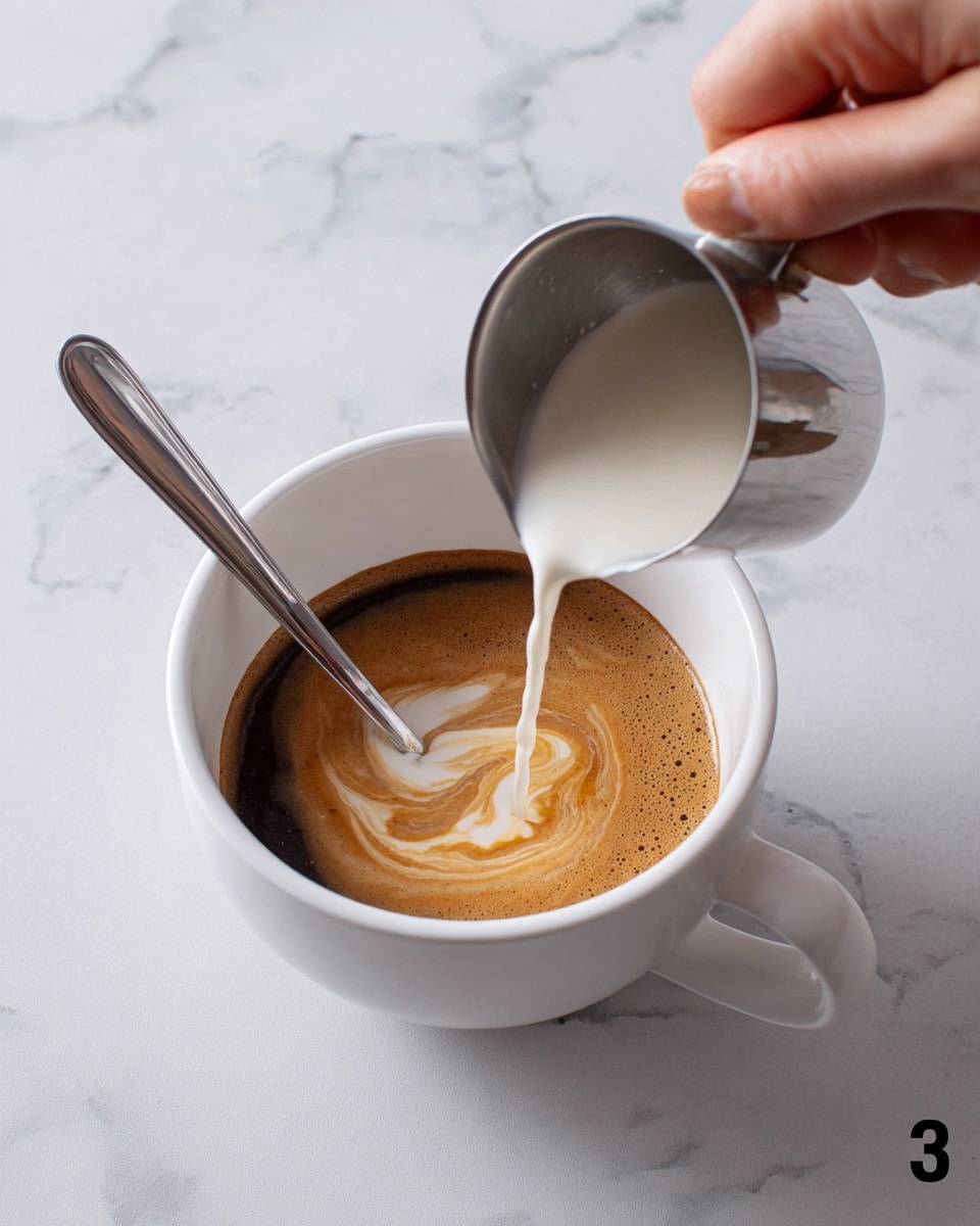 A white cup filled with dark brown coffee has a thin silver spoon resting inside on the left edge. A woman's hand pours a thick layer of light cream from a small shiny metal pitcher into the coffee, creating a smooth, swirling mix of light cream on top of the dark brown base. The cup is on a white marbled surface. photo taken with an iphone --ar 4:5 --v 7
