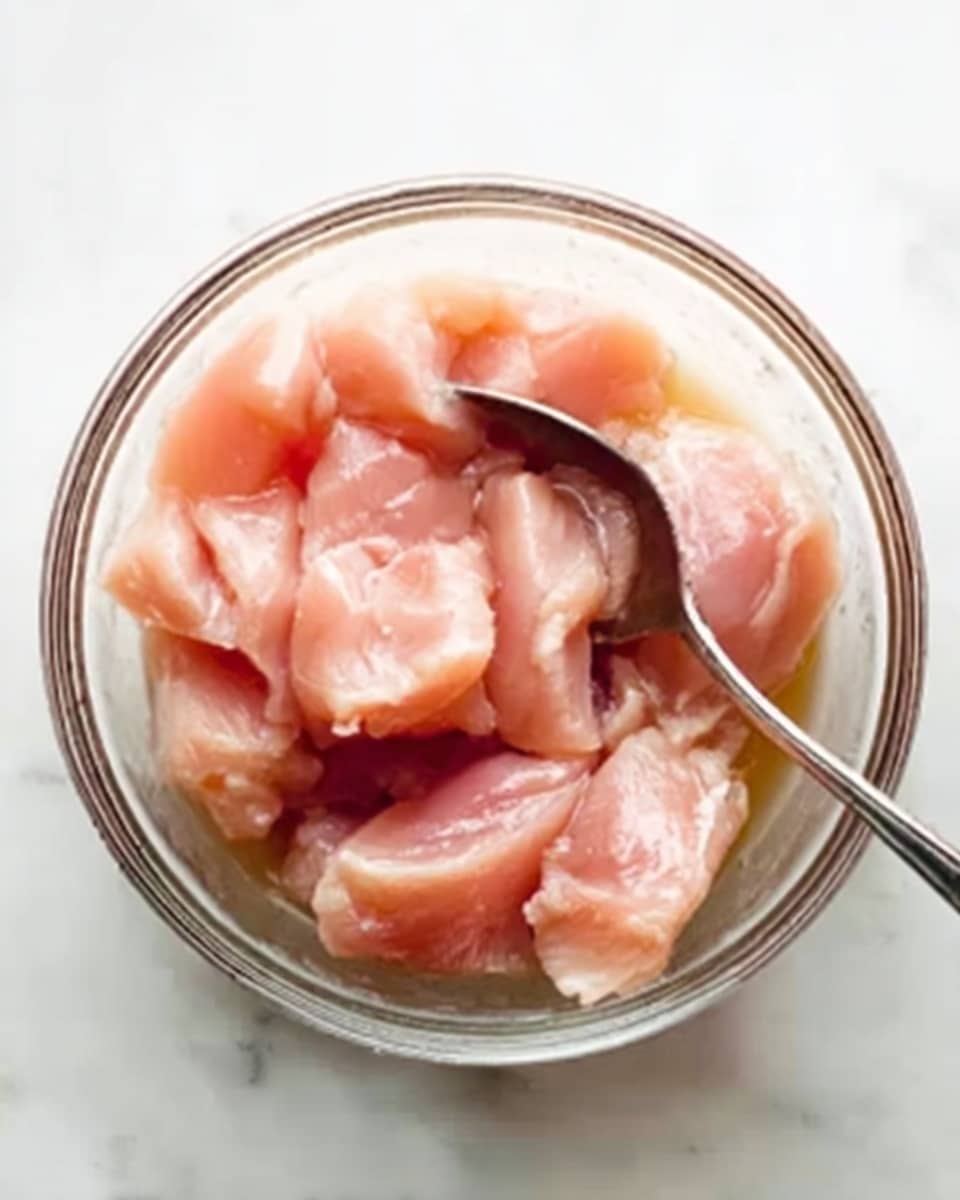 The image shows a clear glass bowl filled with raw light pink chicken pieces that have a smooth, slightly shiny texture. A silver spoon is partially inside the bowl, scooping or stirring the chicken. The bowl is placed on a white marbled surface. Photo taken with an iphone --ar 4:5 --v 7
