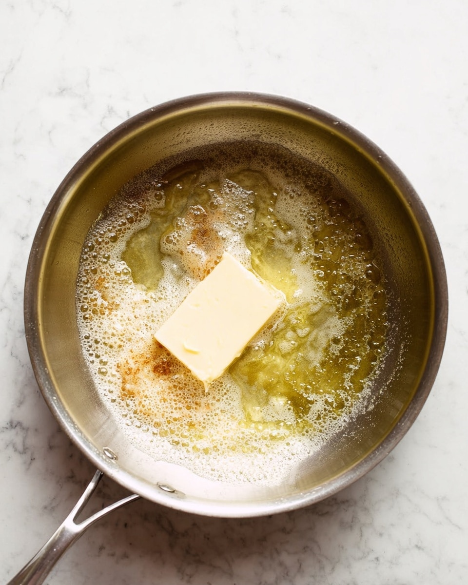 The image shows a silver cooking pan with a melting pale yellow square of butter inside. The butter is partially melted, surrounded by frothy, light golden liquid with some brown patches mixed in, indicating the early stage of browning. The pan has a simple metal handle and sits on a surface with a white marbled texture. The light reflects softly on the bubbling butter and the pan's smooth interior. photo taken with an iphone --ar 4:5 --v 7