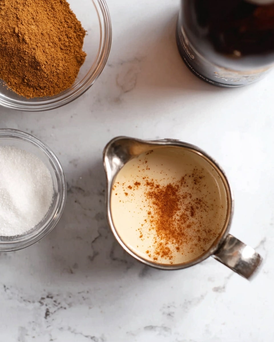A small silver metal jug filled with creamy light brown liquid sprinkled with cinnamon on top sits on a white marbled surface. To the left, there are two small clear glass bowls, one filled with brown cinnamon powder and the other with white sugar. A dark brown bottle is partially visible in the top right corner. The whole scene is shot from above, showing simple textures and warm colors. Photo taken with an iphone --ar 4:5 --v 7