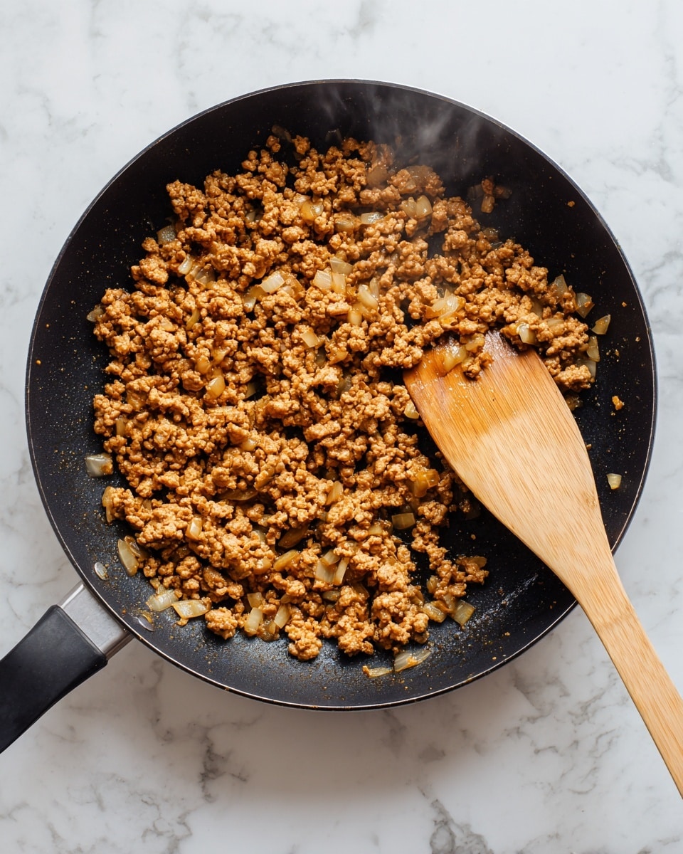 In a black frying pan, there is cooked ground meat mixed with small pieces of light-colored onions, evenly spread out across the pan. A wooden spatula with a smooth, light brown texture rests on the right side, partially lifting some of the meat and onions. The meat has a crumbly texture with a warm brown color, and steam rises gently from the hot mixture. The background surface has a white marbled texture. photo taken with an iphone --ar 4:5 --v 7