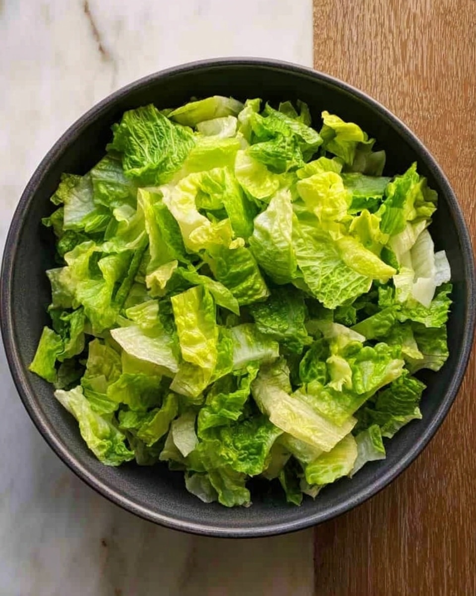 A dark gray bowl filled with a single layer of chopped fresh lettuce leaves in different shades of green, ranging from light to medium green, resting on a white marbled surface. The lettuce pieces have a crisp texture and uneven edges, loosely piled in the bowl. Photo taken with an iphone --ar 4:5 --v 7