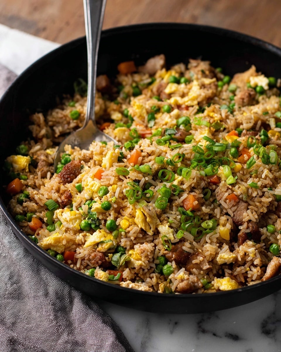A black pan filled with fried rice showing many layers and colors: cooked brownish rice forms the base mixed with small pieces of light yellow scrambled eggs, green peas, bright yellow corn, orange carrot bits, and small brown meat chunks scattered throughout. Green onion slices are sprinkled on top, adding fresh green color. A silver spoon stands upright in the pan. The pan rests on a white marbled surface with a gray cloth visible on the left side. Photo taken with an iphone --ar 4:5 --v 7
