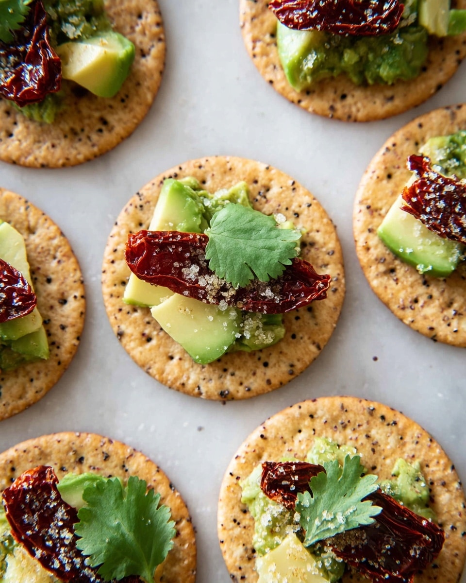 Several light brown round crackers with speckles are arranged on a white marbled surface. Each cracker has a layer of sliced pale green avocado pieces, topped with dark red sun-dried tomatoes that have a wrinkled texture. A small bright green cilantro leaf sits on top of the tomatoes on each cracker, and there is a light sprinkling of coarse salt over the toppings. The image shows close-up details of the layers and textures of each cracker bite, with natural light highlighting the different colors and freshness of the ingredients. photo taken with an iphone --ar 4:5 --v 7