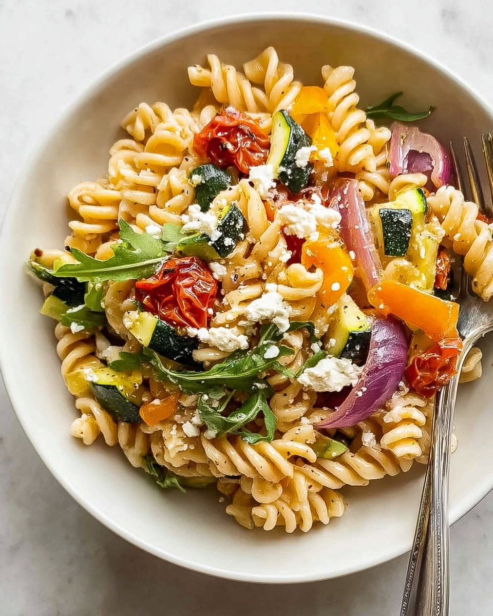 A white bowl filled with spiraled pasta mixed with colorful vegetables and cheese. The pasta is light beige and sits as the base layer, topped with chunks of green zucchini, orange bell pepper pieces, and slices of purple-red onion scattered throughout. Bright red roasted cherry tomatoes and small white cheese crumbles are spread on the top, along with some dark green leafy arugula for a fresh touch. The bowl rests on a white marbled surface next to a silver fork. photo taken with an iphone --ar 4:5 --v 7