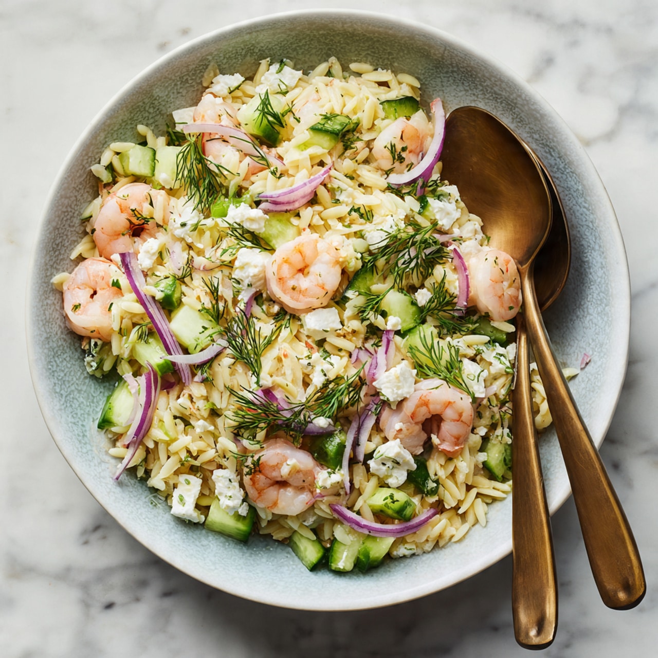A bowl filled with orzo pasta salad layered with pale pink cooked shrimp, small cubes of white cheese, and light green cucumber pieces. Scattered on top are finely chopped red onions, thin green scallion slices, fresh dill sprigs, and flat parsley leaves, all adding green and purple contrast. The bowl is white with a slight blue-gray texture, and two bronze spoons rest inside the bowl on the right side. The background is a white marble surface. Photo taken with an iphone --ar 4:5 --v 7