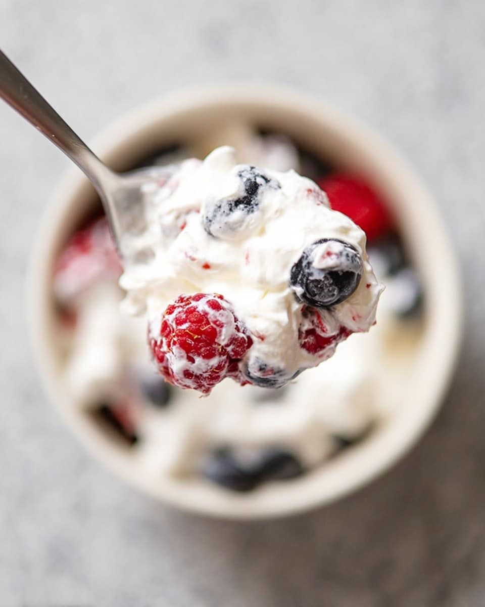 A close-up of a spoon holding a mix of white whipped cream and different berries, including red raspberries with their bumpy texture and dark blueberries, showing a soft and airy texture. The spoon is held above a white bowl filled with the same mixture, which sits on a white marbled surface. The contrast between the creamy white layer and the bright red and dark blue fruits is clear, with the creamy texture wrapping around the berries. photo taken with an iphone --ar 4:5 --v 7