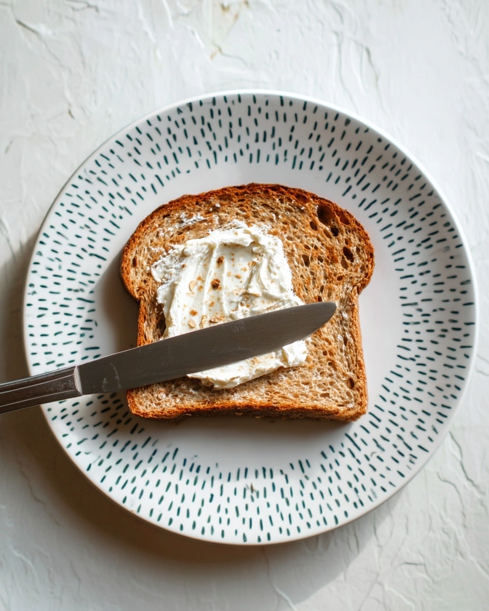 A single slice of toasted whole grain bread with a golden brown color and visible seeds and grains is placed in the center of a white plate with small blue dotted circular patterns. On top of the bread, there is a thick layer of white creamy spread being smoothed with a silver knife, which is positioned diagonally across the toast. The scene is set on a white marbled surface with soft natural light highlighting the textures of the bread and cream. photo taken with an iphone --ar 4:5 --v 7