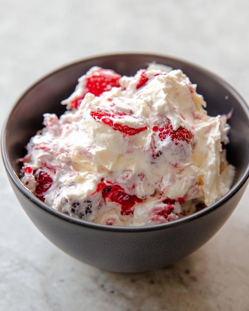 A close-up of a dark gray bowl filled with a creamy mixture containing visible layers of bright red raspberries and white whipped cream, with some parts looking fluffy and soft, creating a textured, mixed look. The bowl is placed on a white marbled surface. The cream covers and blends with the raspberry pieces on top and inside the bowl, showing some shadow and light reflections on the cream’s swirls and berry surfaces. Photo taken with an iphone --ar 4:5 --v 7