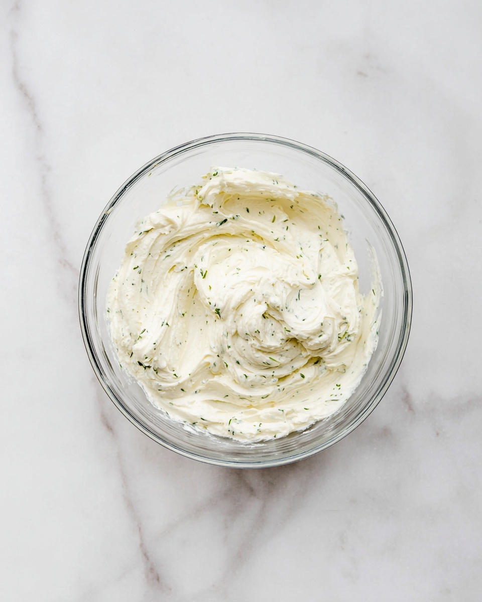 A clear glass bowl sits on a white marbled surface, filled with one thick layer of creamy white mixture that has small green herb specks evenly spread throughout. The texture looks smooth and soft with visible folds and swirls on the top, showing how it was mixed or stirred. The bowl is viewed from above, centered in the image, with the white marbled background giving a clean, bright look photo taken with an iphone --ar 4:5 --v 7