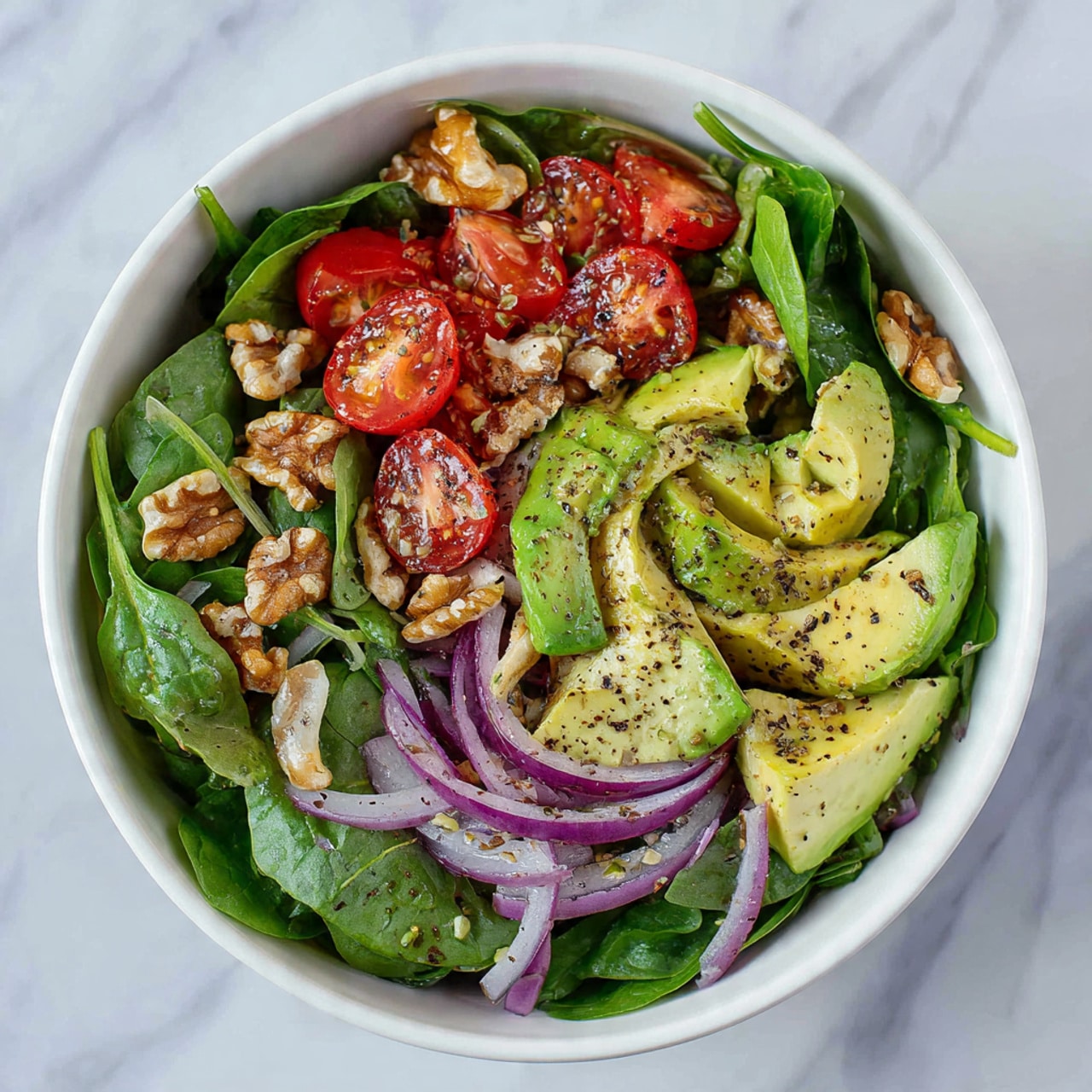A white bowl filled with a fresh salad showing three main layers: the bottom layer is bright green leafy spinach, the middle layer has small bright red cherry tomato halves and purple onion slices, and the top layer is light green avocado chunks mixed with brown walnut pieces, all sprinkled with black pepper. The bowl is placed on a white marbled surface. photo taken with an iphone --ar 4:5 --v 7