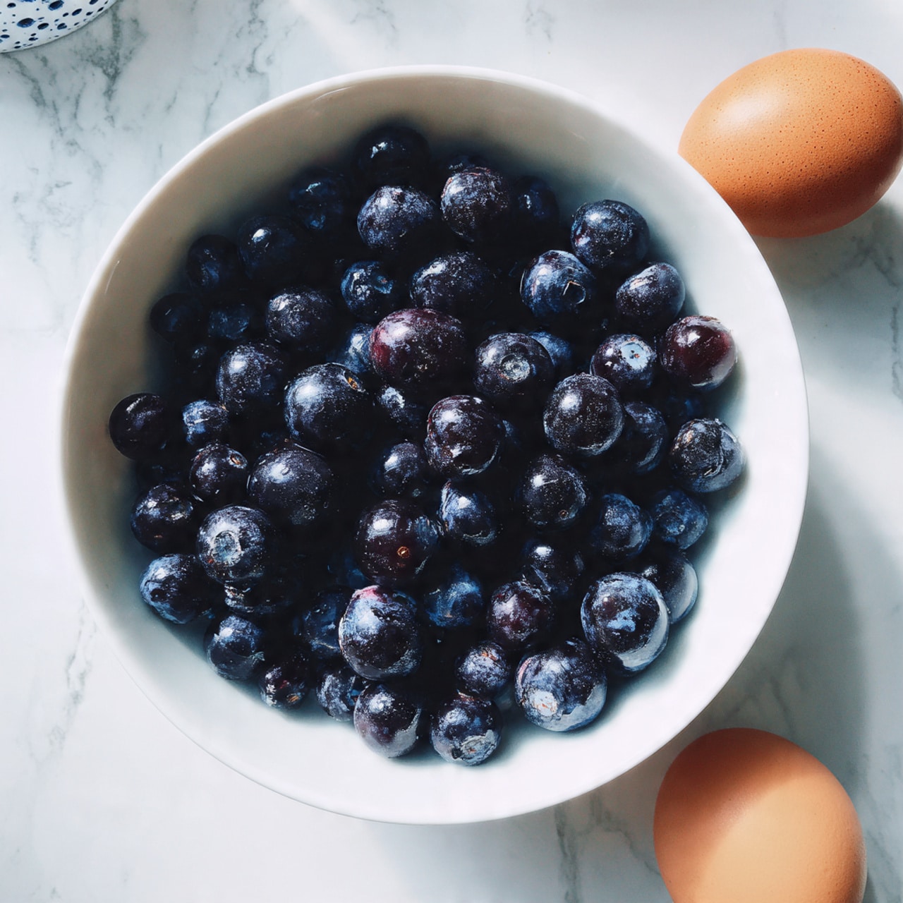 A white bowl filled with many small, round blueberries, dark blue to almost black in color, with a slight natural shine and a few showing light white powdery bloom. The bowl is placed on a white marbled surface. At the bottom right corner, there is a small part of a single brown egg visible. Photo taken with an iphone --ar 4:5 --v 7