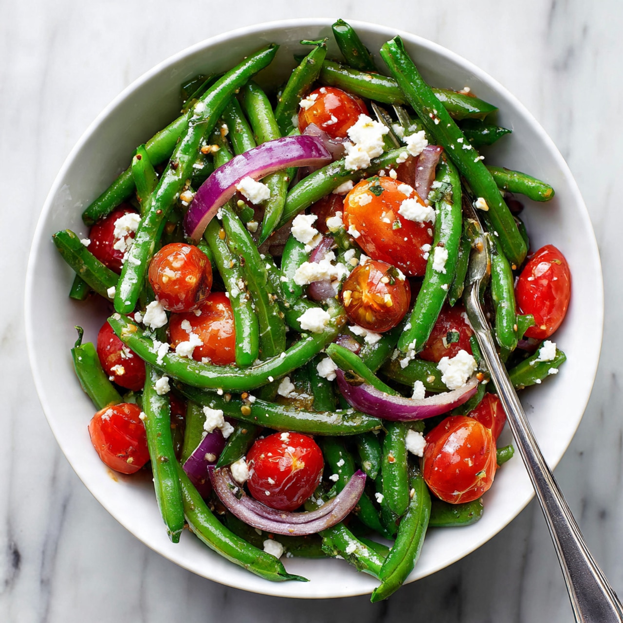 The image shows a close-up of a fresh salad in a white bowl with a white marbled texture beneath it. The salad has three main layers: the bottom layer has cut green beans with a smooth, firm texture; the middle layer has halved bright red cherry tomatoes with a shiny, juicy look; the top layer has crumbled white feta cheese scattered unevenly, along with thin slices of purple onions adding a slightly translucent texture. There is a light, glossy dressing drizzled over the salad, giving it a fresh and appetizing look. photo taken with an iphone --ar 4:5 --v 7
