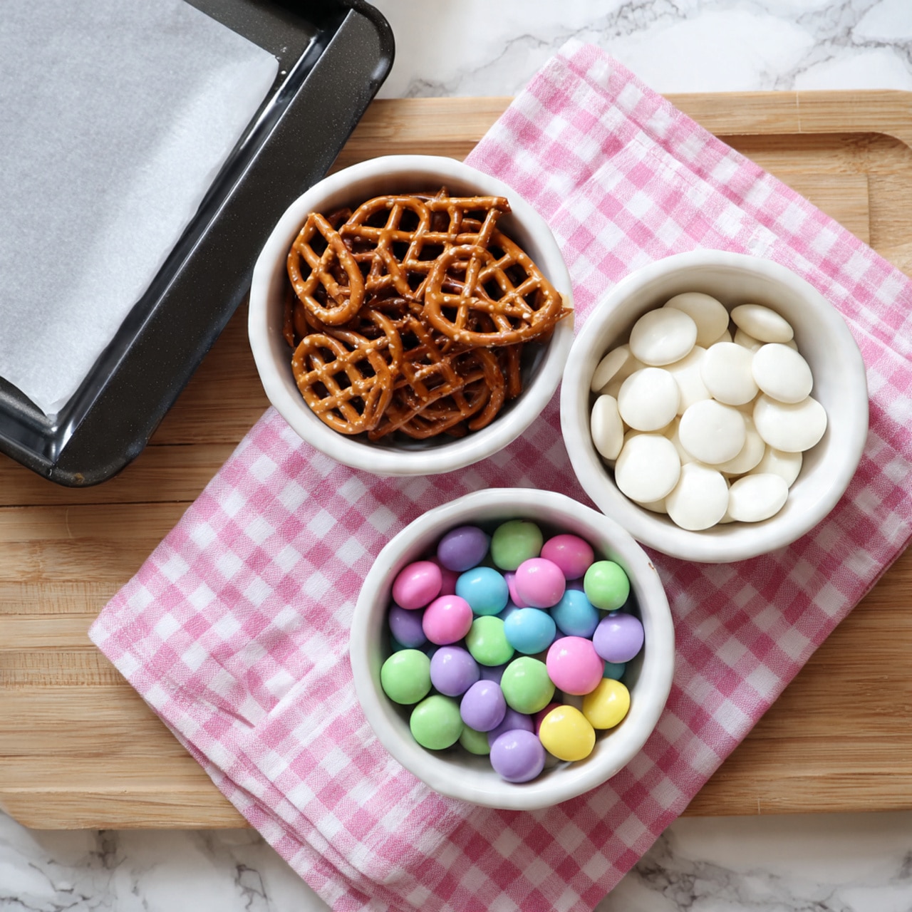 The image shows three small white bowls placed on a pink checkered cloth over a wooden surface. One bowl is filled with small, square-shaped brown pretzels with a grid pattern. The second bowl contains round, flat white candy melts with a smooth texture. The third bowl holds colorful round candy-coated chocolates in pastel shades of purple, pink, green, yellow, and blue. To the left of the bowls, there is a black baking tray lined with white parchment paper resting on the wooden surface. The whole scene is bright and neatly arranged, with a white marbled texture in the background. photo taken with an iphone --ar 4:5 --v 7