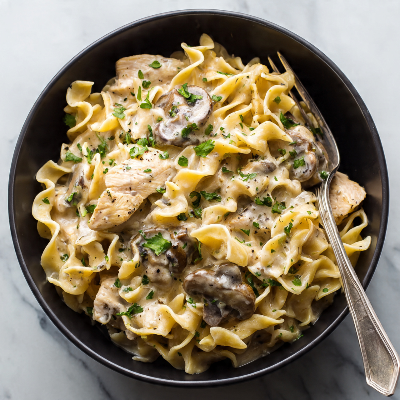 A black bowl holds wide egg noodles mixed with light brown cooked chicken slices and dark brown mushroom pieces, all coated in a creamy white sauce. The noodles have a soft, slightly curled texture and the chicken pieces are tender, scattered evenly throughout. Small green parsley leaves are sprinkled on top for color, and a silver fork lies on the side inside the bowl. The bowl sits on a white marbled surface. photo taken with an iphone --ar 4:5 --v 7